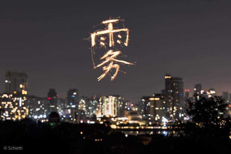 A city skyline at night with blurred lights; above the buildings, glowing Chinese characters appear as if drawn with light, creating a striking contrast against the dark sky.