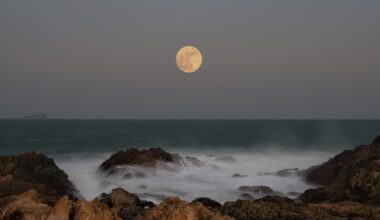 Photo of a full moon over the ocean, taken from rocky shore. Waves can be seen crashing over the rocks in the foreground/bottom of the image.