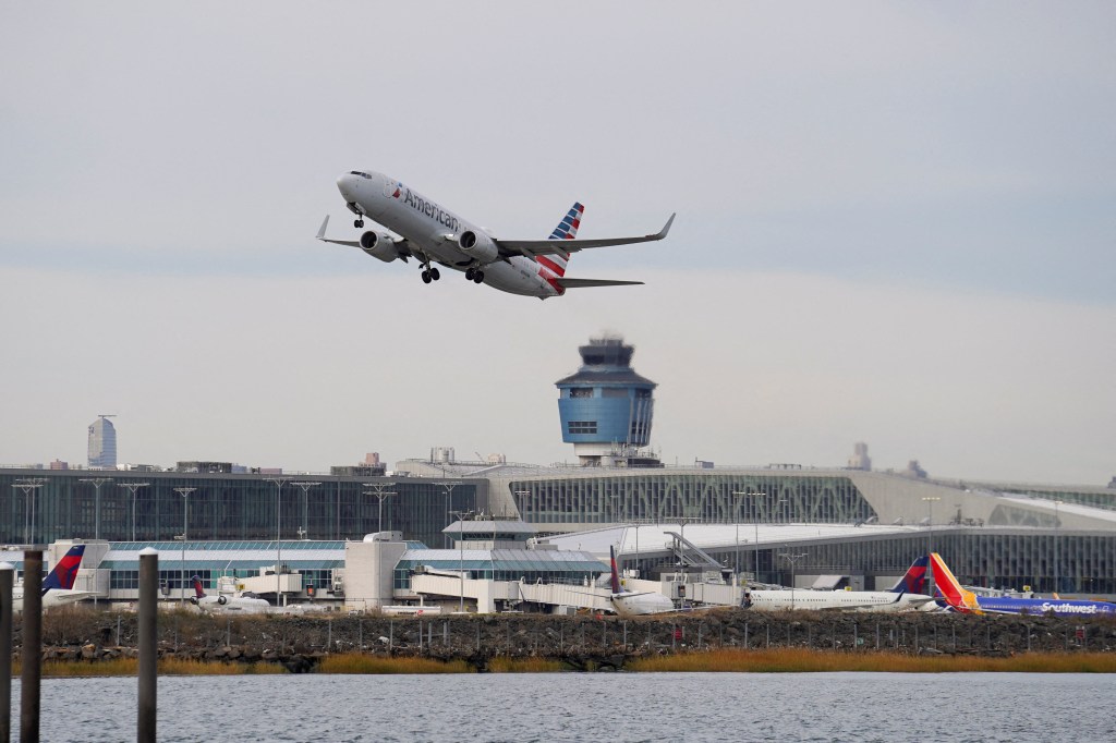 An American Airlines plane takes off from LaGuardia Airport over a body of water, with the air traffic control tower and airport terminals visible.