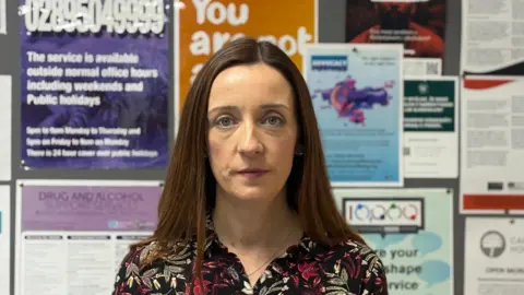 Katherine Kidd has long brown hair. She is wearing a black shirt with pink and green floral patterns. She has a thin chain necklace around her neck. She is standing in front of a notice board with lots of coloured posters on it.
