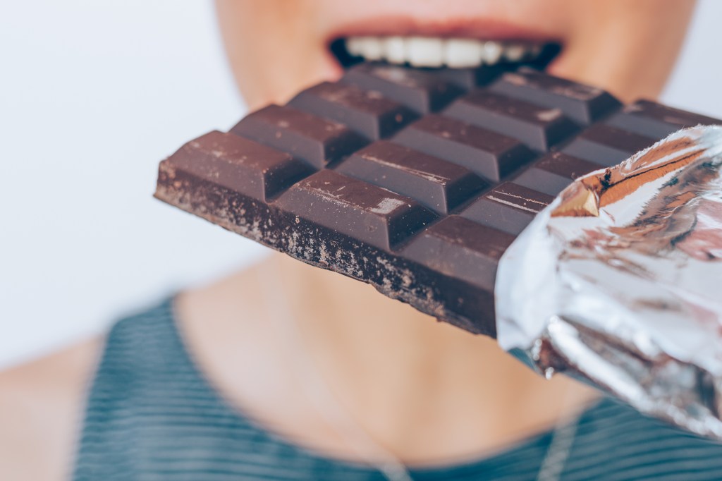 A young woman biting into a large dark chocolate bar, with the foil wrapper still visible on the right side.