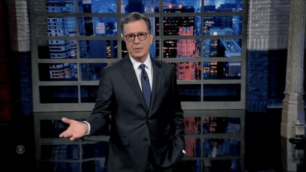 Stephen Colbert in a suit gesturing with his left hand in front of a city skyline backdrop.