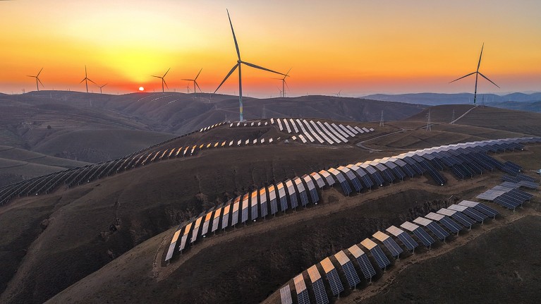 An aerial view of the sun setting behind wind turbines and rows of solar panels at a wind farm in China.