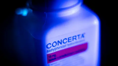 Getty Images A bottle of Concerta pills ADHD medication. The bottle is white but is lit up blue. The background is black. 