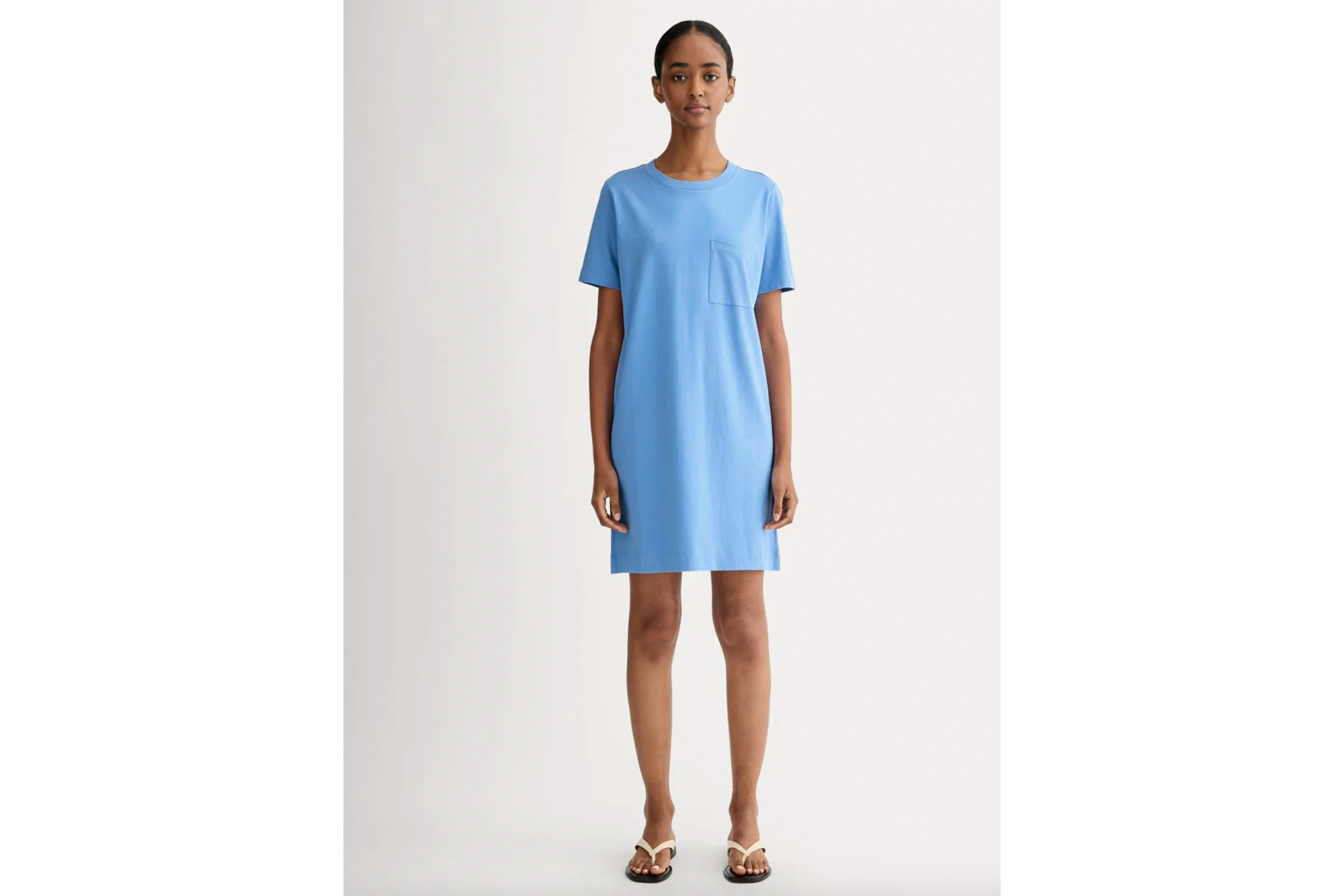 Woman wearing a light blue t-shirt dress with a chest pocket and white flip-flops.