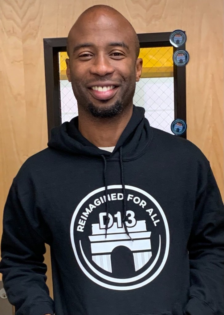 District Superintendent Kamar Samuels smiling and wearing a black hoodie with "REIMAGINED FOR ALL D13" logo.