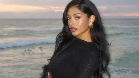 Kaaviya Sambasivam Sambasivam wears a black top and gold earrings while standing in front of the ocean