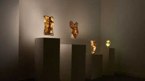 BETHLEM GALLERY Four stands, there are four gold coloured abstract sculptures which are lit from below.