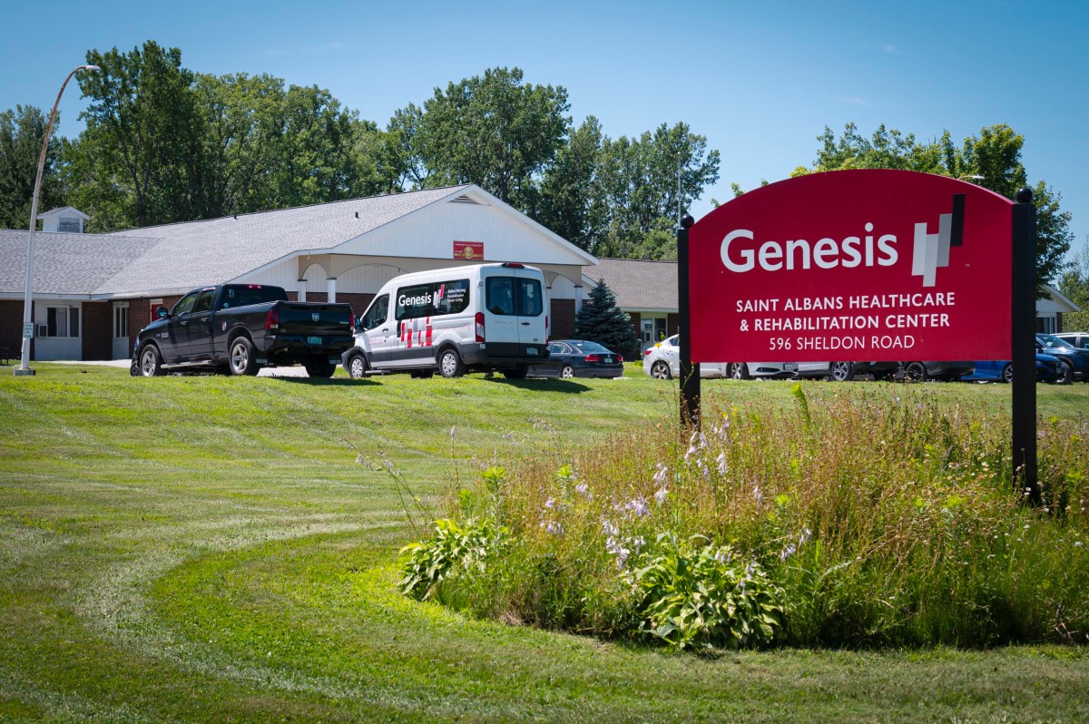 A red sign for Genesis Saint Albans Healthcare & Rehabilitation Center stands in front of a building and parked vehicles on a grassy lot.