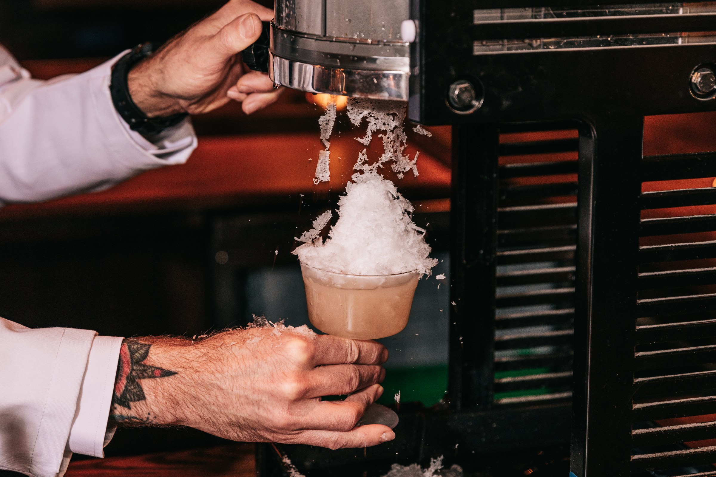 A hand holds a glass under a shaved ice machine.