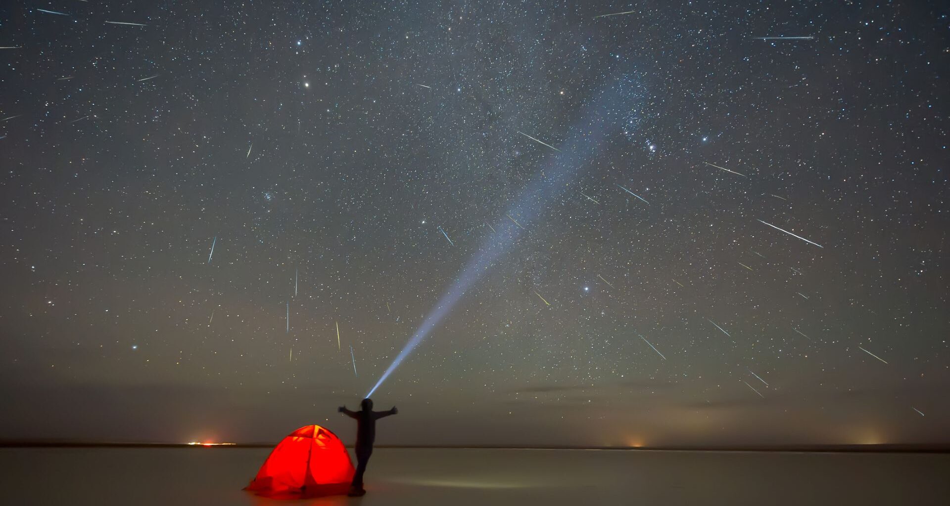 A person stands with arms outstretched behind a bright orange tent looking up at the night sky full of streaks with meteors with a large flashlight beam pointed up.