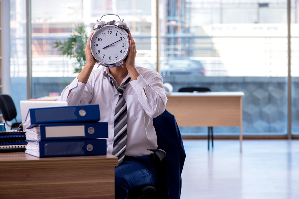 An employee sitting at a desk with his head behind an alarm clock, with stacks of binders and papers next to him, indicating excessive work and stress.