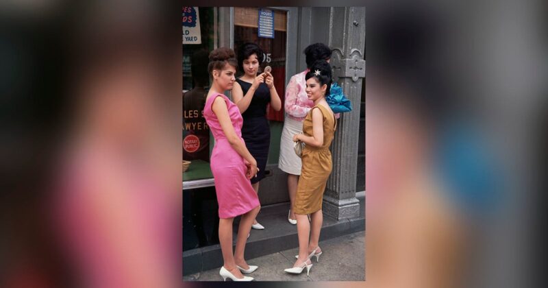 Four women in colorful, form-fitting 1960s dresses and white heels stand on a city sidewalk near a storefront, chatting and posing. Two are facing the camera, while two are turned slightly away.