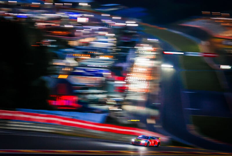 A race car speeds along a track at night, with bright, colorful lights and blurred buildings in the background, creating a sense of motion and excitement.