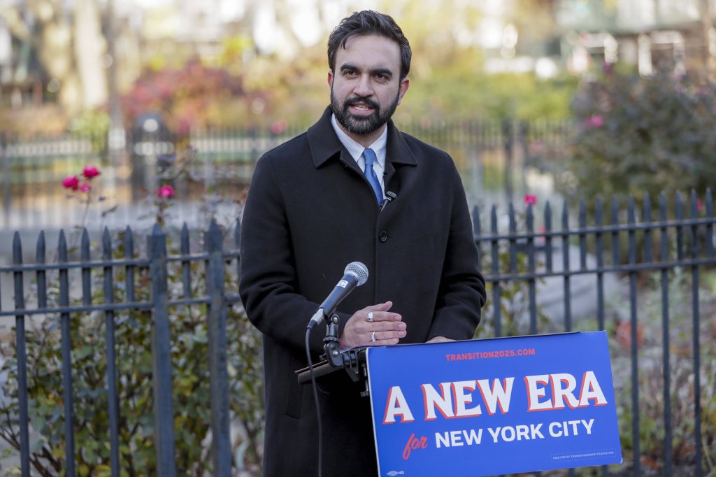 New York City Mayor-elect Zohran Mamdani speaking at a podium with a sign reading "A New Era for New York City."