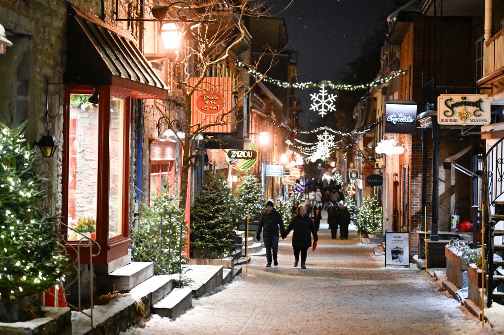 Petit-Champlain street in Quebec City decorated for Christmas with storefronts, string lights, snowflakes, and small evergreen trees, with several people walking on the snow-dusted ground.