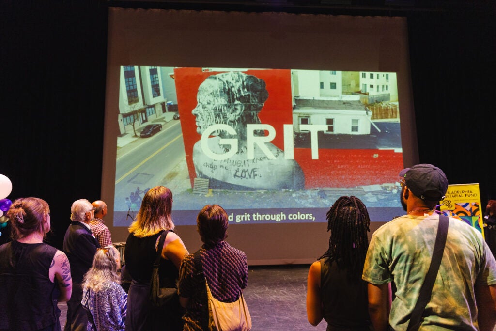 People stand in front of a projected image showing an art installation with the word "GRIT" in front of it