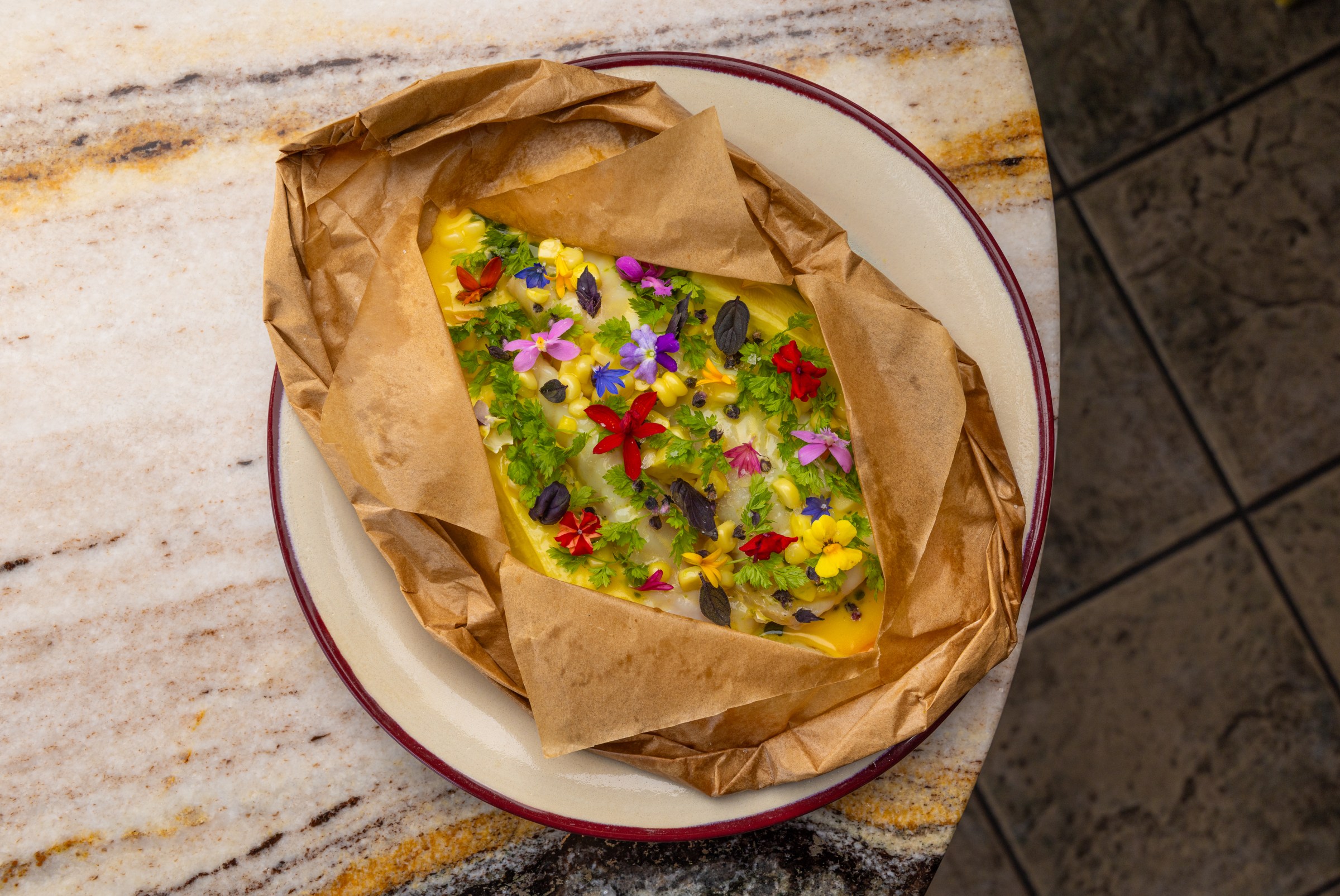 Overhead shot of paper opening up to reveal fish with summer corn, kohlrabi, cipollinis, calabaza, and saffron.