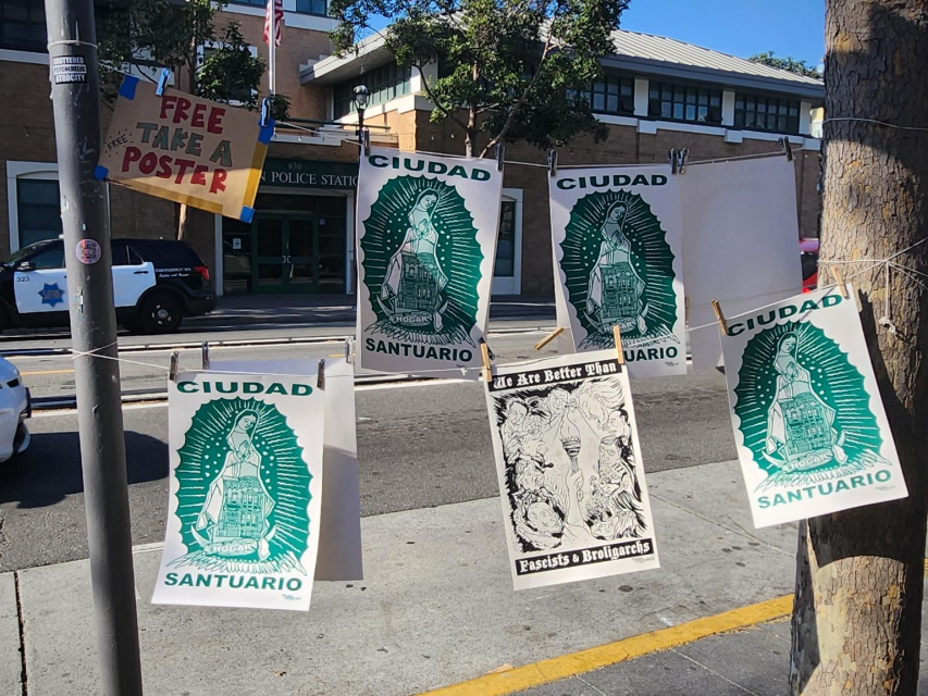 Several "Ciudad Santuario" posters and one black-and-white poster hang on a clothesline near a police station; a sign offers free posters to passersby.