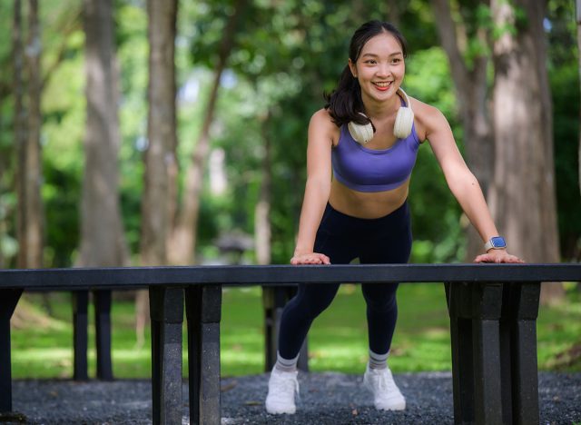 Young Asian woman performing incline push-ups using a park bench, exercising outdoors to stay fit and healthy
