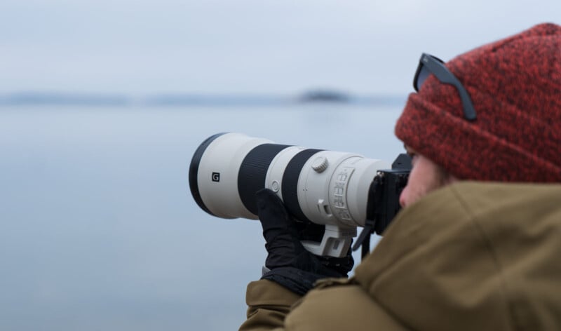 Person in a red beanie and brown jacket using a large camera with a telephoto lens to take a photo of a distant landscape over water. The scene appears cold and overcast.