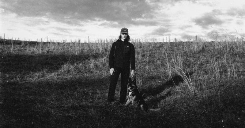 A person stands in a grassy field with a dog sitting at their feet. The image is black and white, with tall grass and a cloudy sky in the background, creating a moody, natural atmosphere.