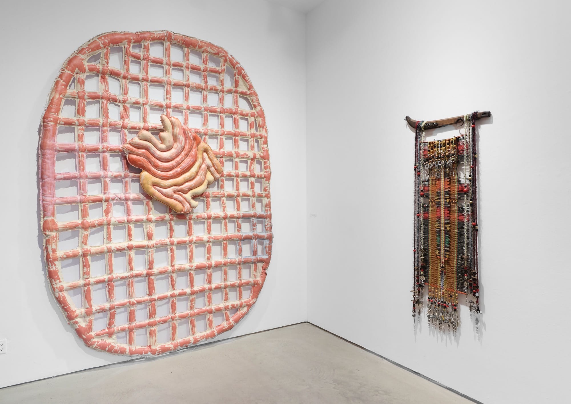 An installation view of 'Thread Count' featuring two wall works made from textiles. The one of the left is a grid pattern within an overall oval shape in orange-pink, and the one on the right is a mixed-media weaving hanging from a piece of wood