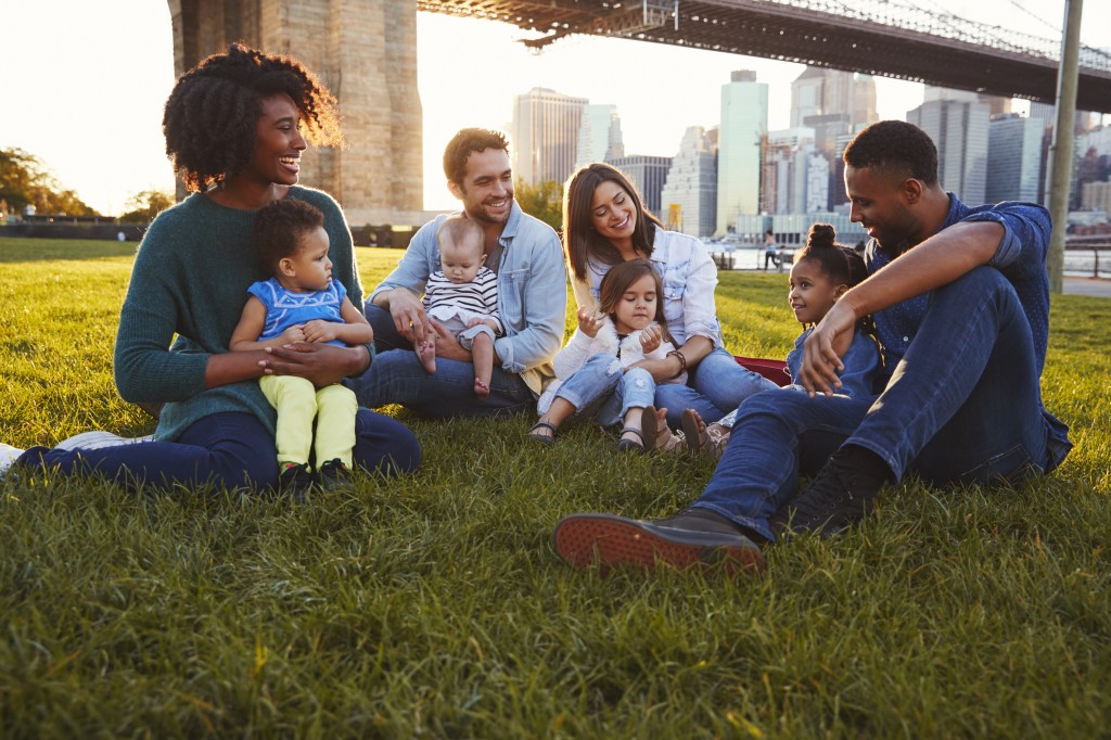 Two families with young daughters and babies sitting on a lawn in Manhattan, New York.