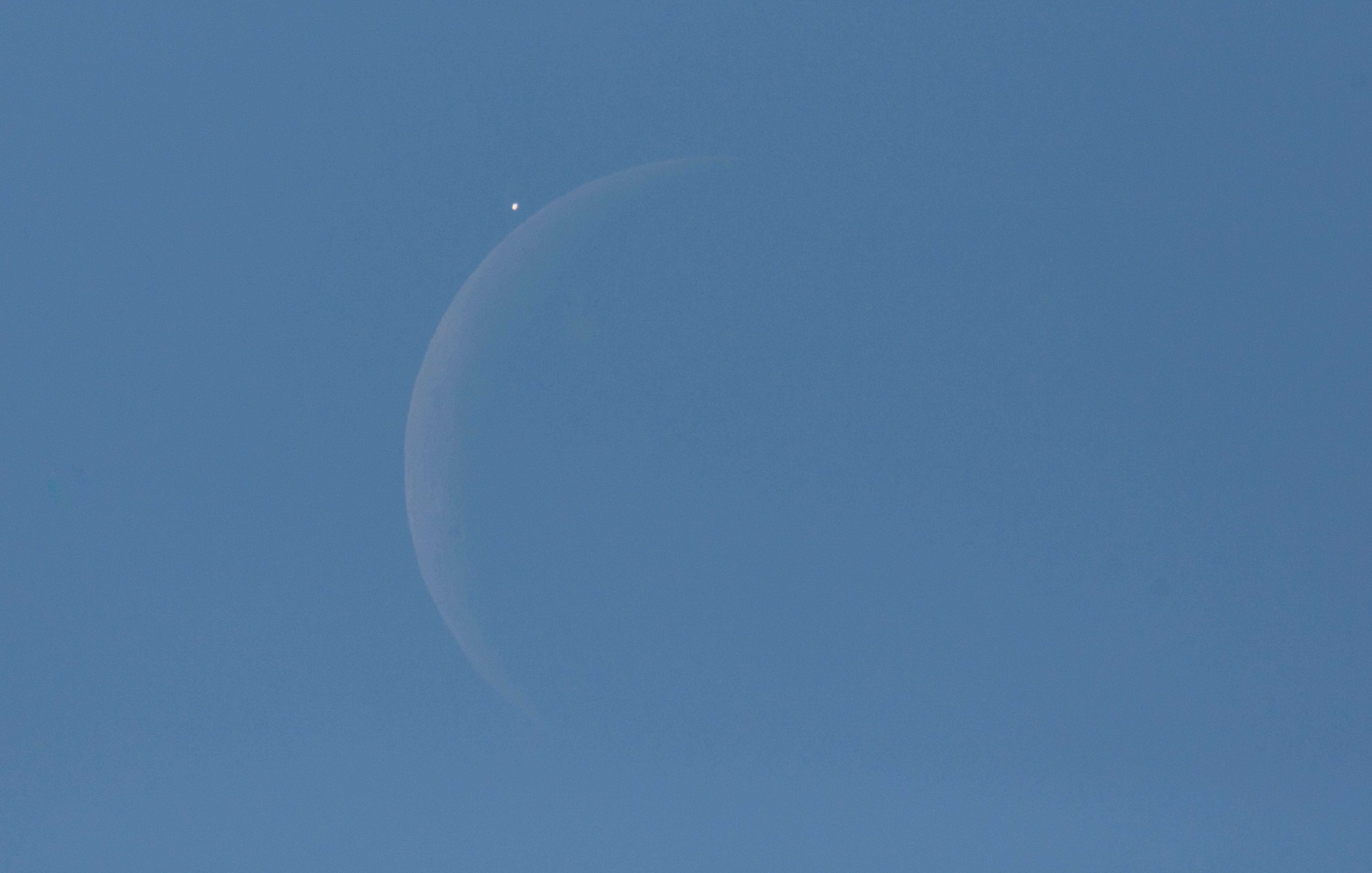 Venus is seen next to the crescent moon during the daytime against a blue sky, prior to the start of occultation.