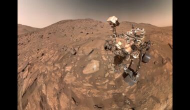 Photo of the Perseverance rover taken by the rover from above. The Martian landscape makes up most of the photo&mdash;reddish brown rocks and dirt.