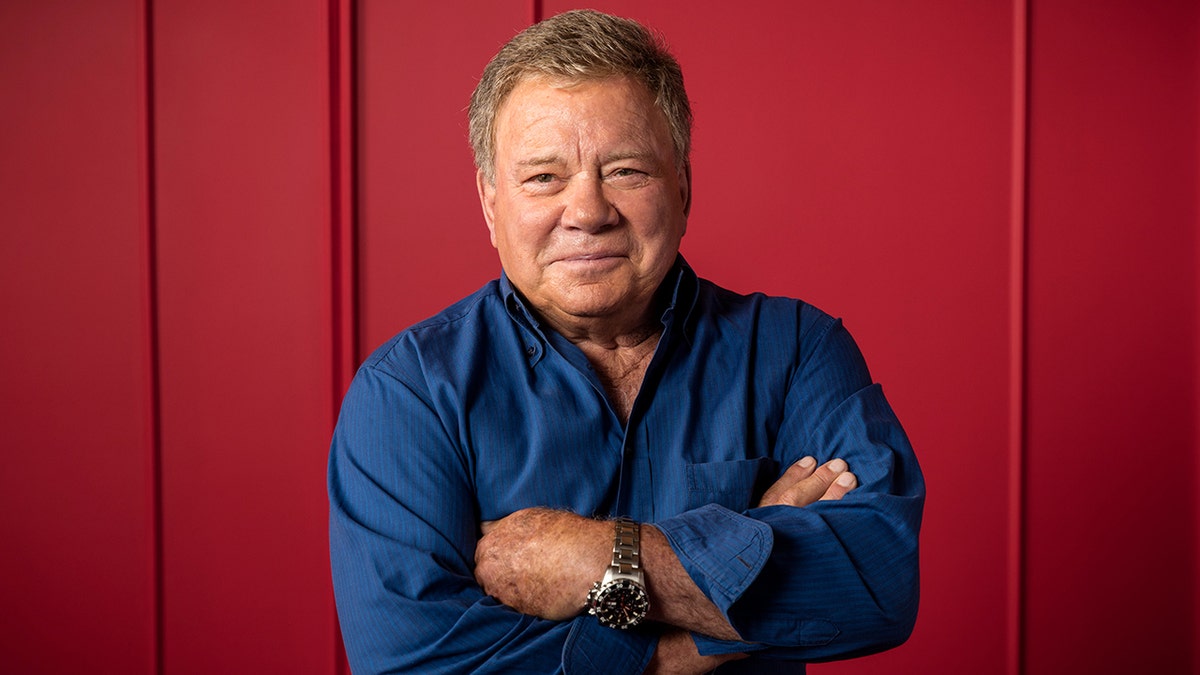 William Shatner in a blue shirt crosses his arms over his chest and soft smiles.