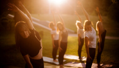 A group of women perform yoga in a park outdoors, each in the same pose, standing with their left arm extended overhead. The sun shine down from the top of the image.