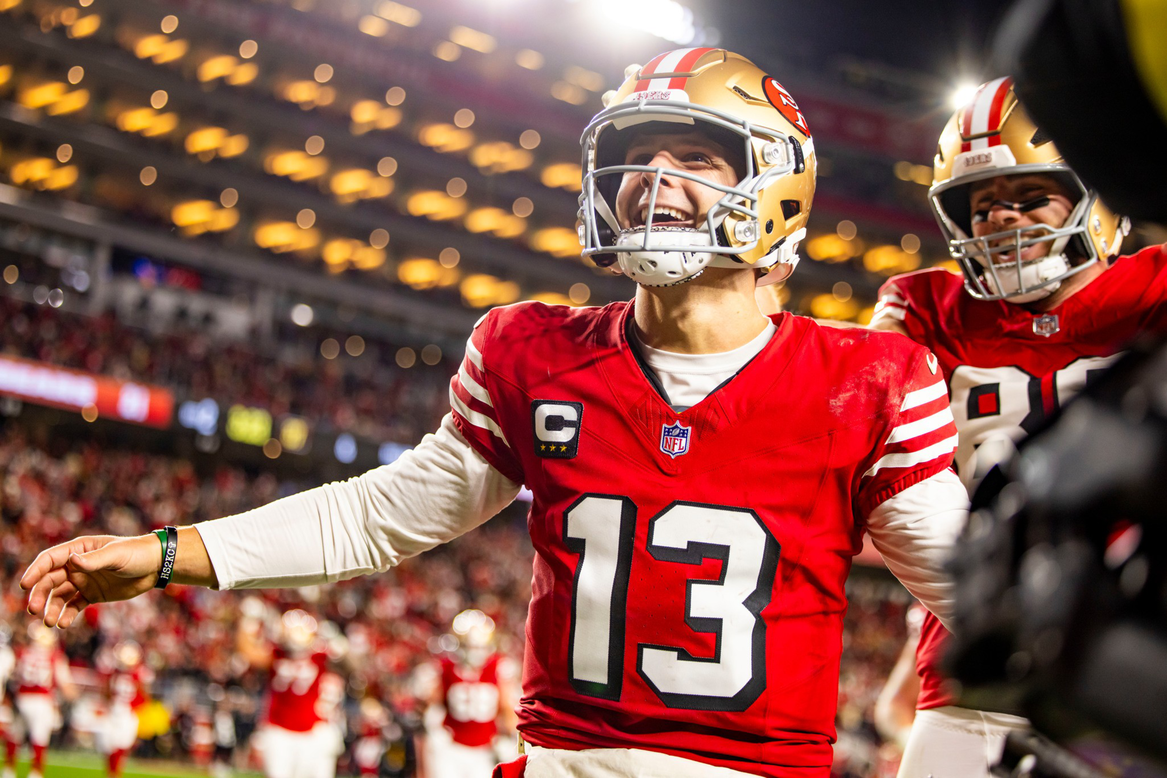 A smiling football player wearing a red number 13 jersey and gold helmet celebrates on the field with a teammate behind him, stadium lights glowing.