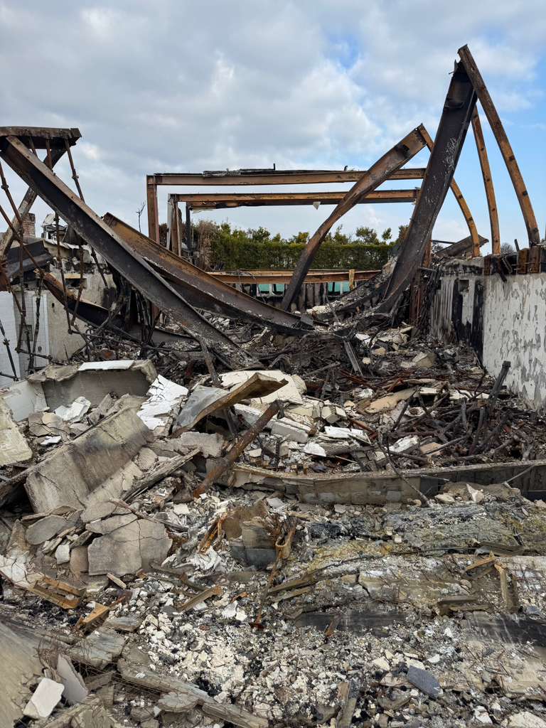 The charred remains of a building after a fire, with twisted metal beams and rubble scattered across the ground.