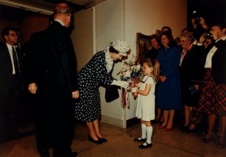 A woman bends to receive flowers from a little girl as well-dressed adults watch them from either side.