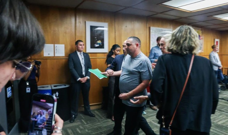 Several people watch as two men escort a third man holding papers in a hallway. A cameraman records the scene in the foreground.