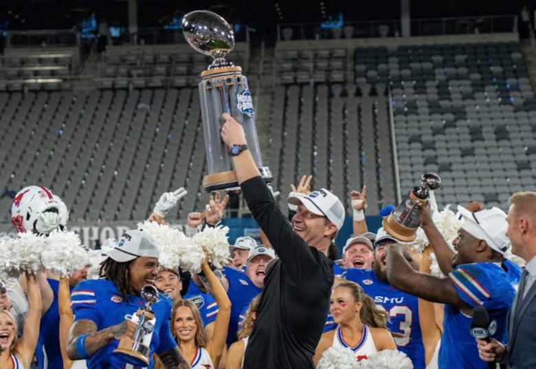 A man surrounded by happy football players raises a trophy above his head against a backdrop of empty stands after the Holiday Bowl in San Diego.