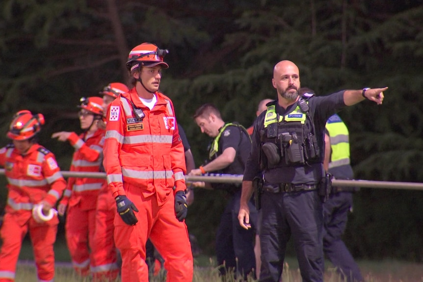 SES officers in orange overalls and helmets stand near a police officer in navy who is pointing off camera.