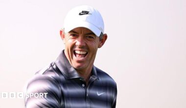 Rory McIlroy appears to be laughing while wearing a golf shirt with a black and white hooped effect, and a white cap