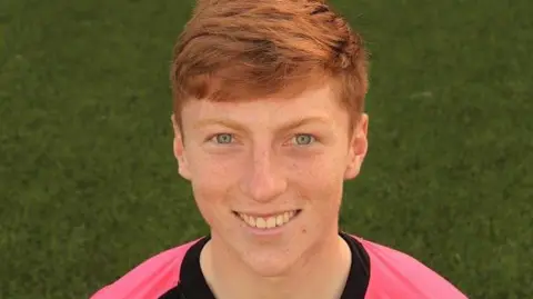 Queen of the South A young man with ginger hair in a pink football top with grass in the background