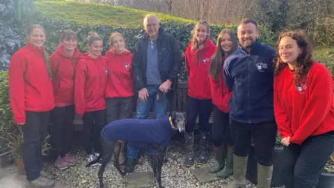 Woodside Animal Welfare Trust Nine people stood together outside with the greyhound dog, Bobby, at the centre. The dog is wearing a blue coat. The volunteers, stood either side of Peter Hart, are wearing red coats. Peter is wearing a black coat. They are all smiling at the camera.