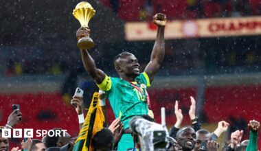 Brahim Diaz in all-white Morocco kit is seen on the left side of an image, chipping a penalty straight into the arms of Senegal goalkeeper Eduoard Mendy, all in blue on the right of the image, in the final of Afcon 2025. Mendy waits, bent slightly over, to collect the ball which is nearing him