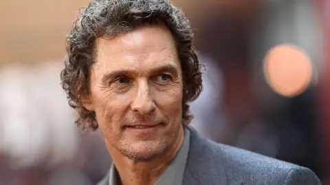 AFP via Getty Images Matthew McConaughey is smiling at a camera. He has short brown curly hair, and is wearing a grey suit. This photo was taken at a screening for a film he stars in called "The Lost Bus”. 