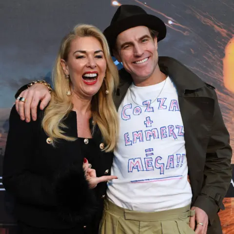 Getty Images Claire Sweeney and James Cartwright smile and stand together in front of Corriedale backdrop at the premiere. He has his arm around her shoulder and he's wearing a white T-shirt with a DIY slogan written in pink and white. She's laughing and pointing at it.