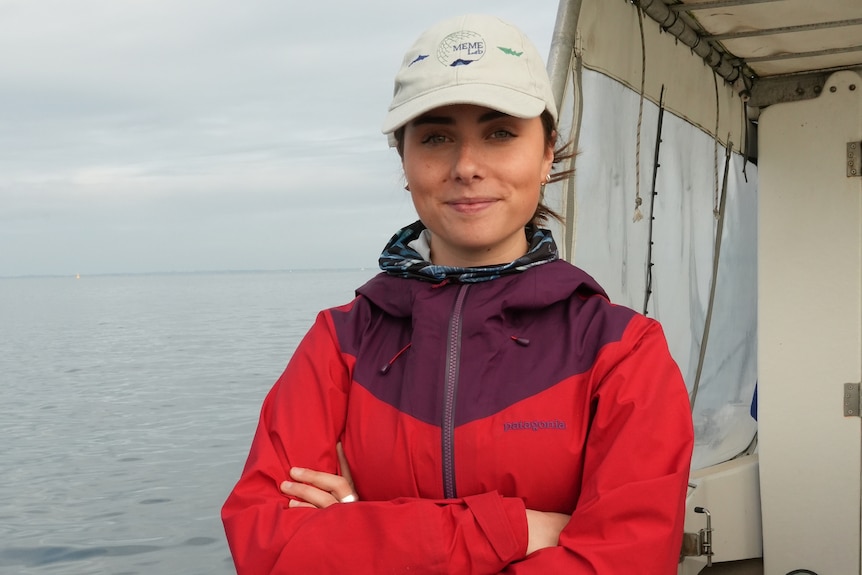 A woman in a red spray jacket with maroon top section and beige cap standing on back of boat with arms crossed.