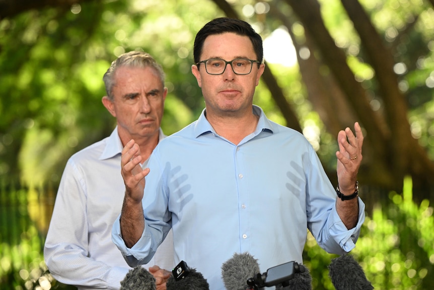 Two middle-aged men in button-up shirts speak to media in front of a leafy green background.