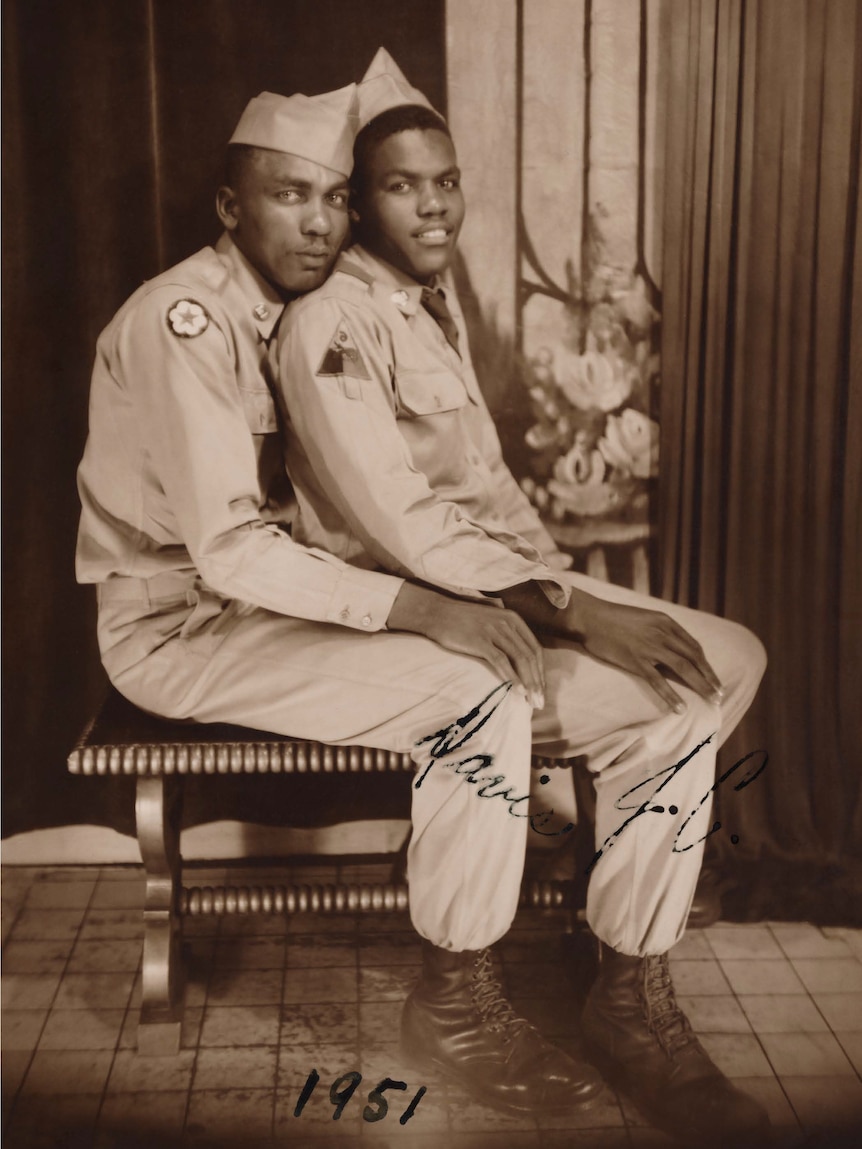 Two men in uniform sit hugging on a bench seat. The photo has the date 1951.