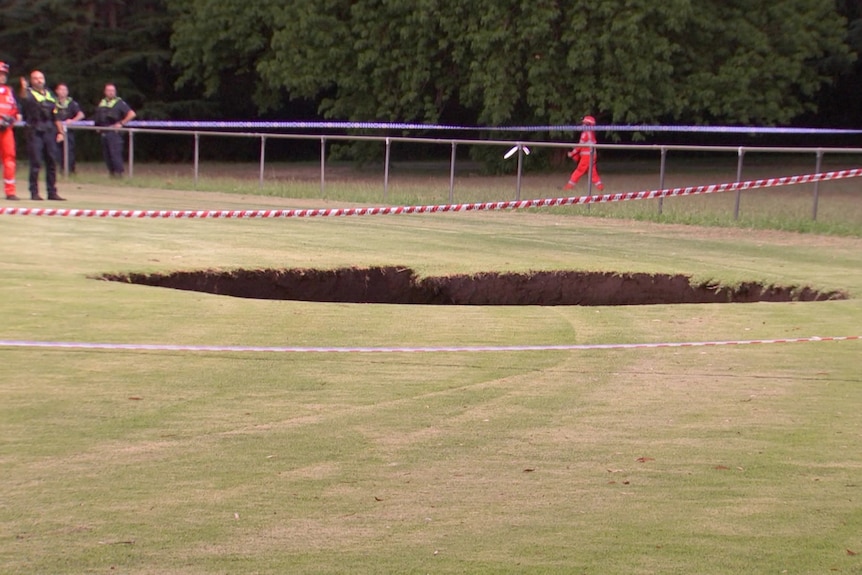 An SES worker in orange overalls and helmet and a police officer stand near a large round hole in the green grass.