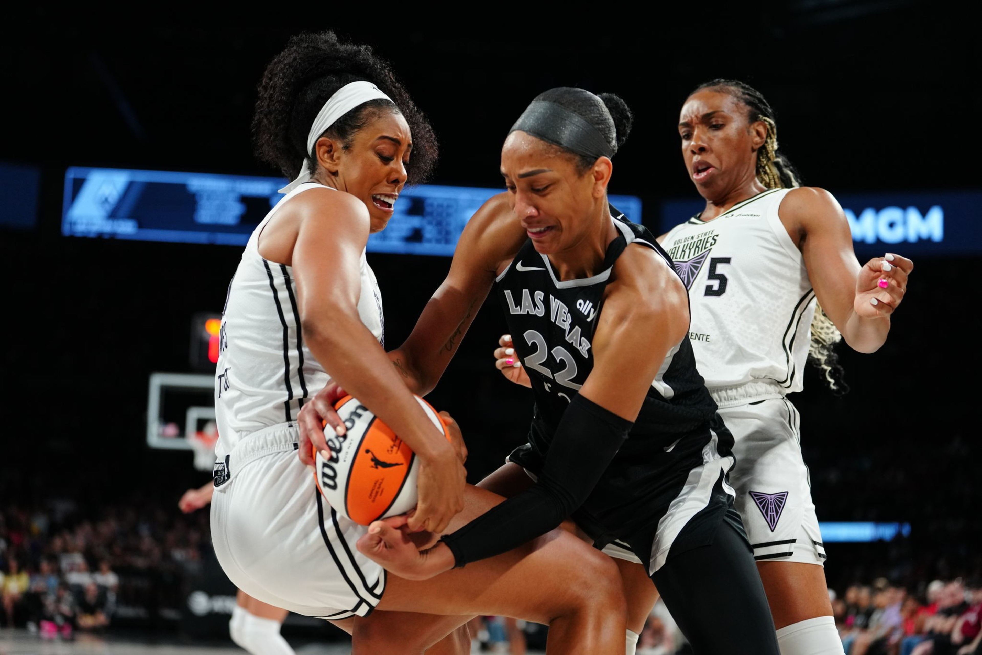 Two basketball players are grasping a ball, each wearing contrasting uniforms, while a third player in a white uniform watches. The scene is intense and dynamic.