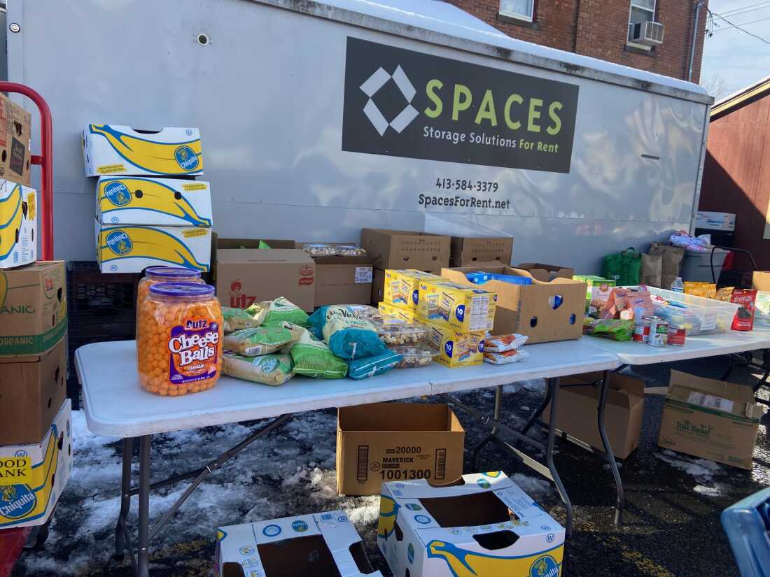 This photo shows a pop-up food bank set up outdoors outside a community center. Groceries are arranged on two folding tables. The food includes canned goods, puffed cheese balls and other nonperishable groceries in boxes and bags. Cardboard boxes are stacked around the tables.